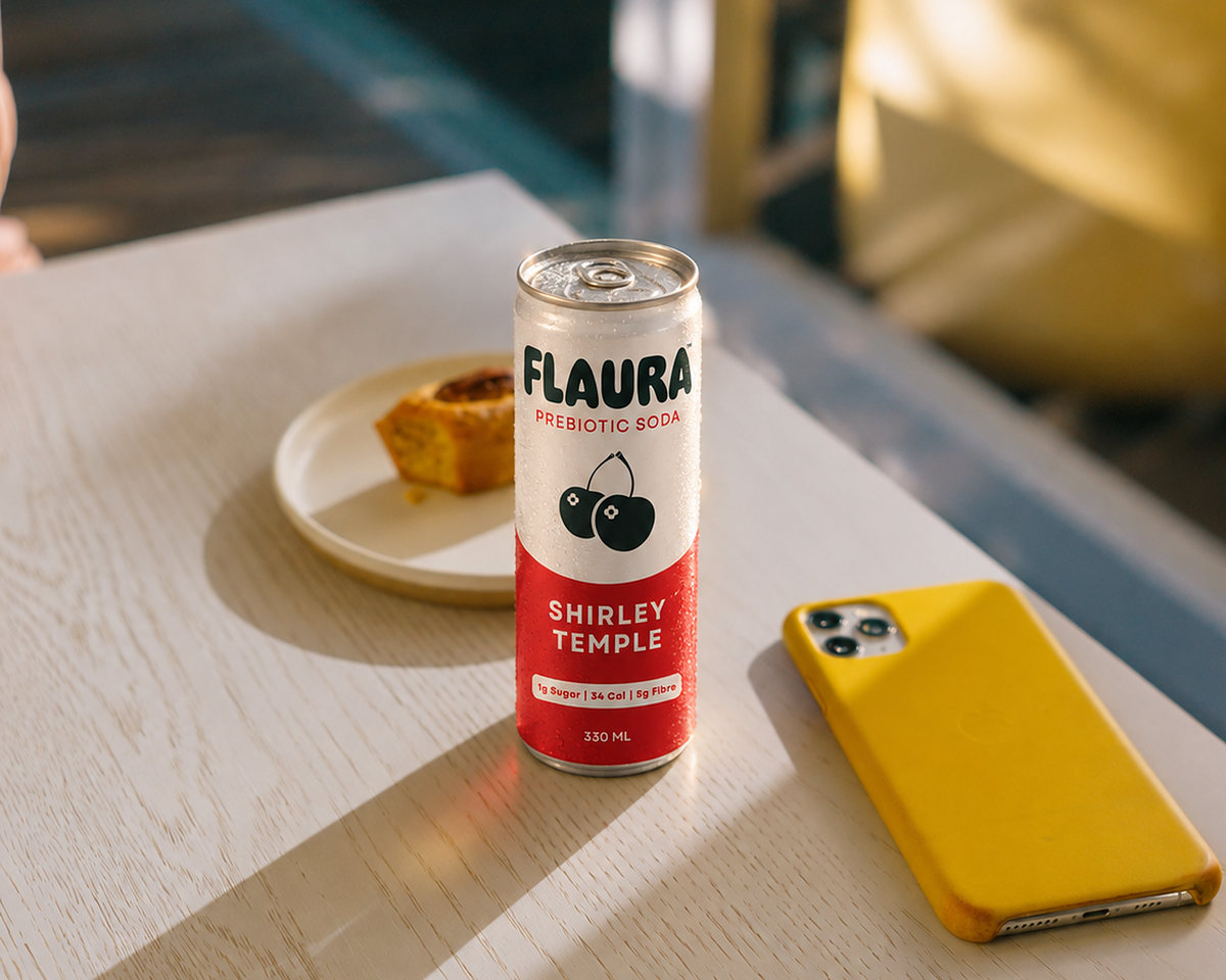 A can of FLAURA Shirley Temple prebiotic soda stands on a light wood table in a real environment, with a small plate, a yellow phone case, and a blurred chair in the background.