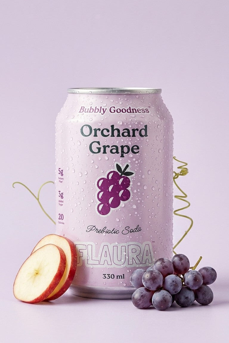 A can of Bubbly Goodness Orchard Grape Probiotic Soda is shown on a light lavender background, styled with a sliced apple and a cluster of purple grapes. The can features a white body with purple and green grape graphics, and the brand name 'FLAURA' is visible at the bottom.