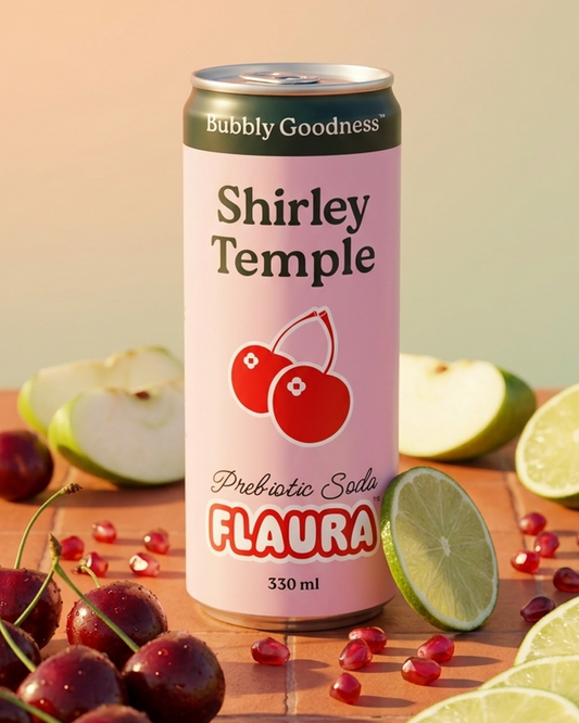 A 330 ml can of Shirley Temple Flaura Prebiotic Solo soda is displayed on a terracotta surface with fresh cherries, lime slices, and pomegranate seeds. The can features a light pink label with a red cherry illustration and a dark green top band reading 'Bubbly Goodness'.