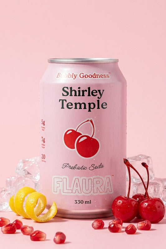 A 330 ml can of Shirley Temple Probiotic Seltzer by Flaura, presented in a pink can with red cherries on the label, surrounded by fresh cherries, pomegranate seeds, and a lemon twist on a solid pink background.